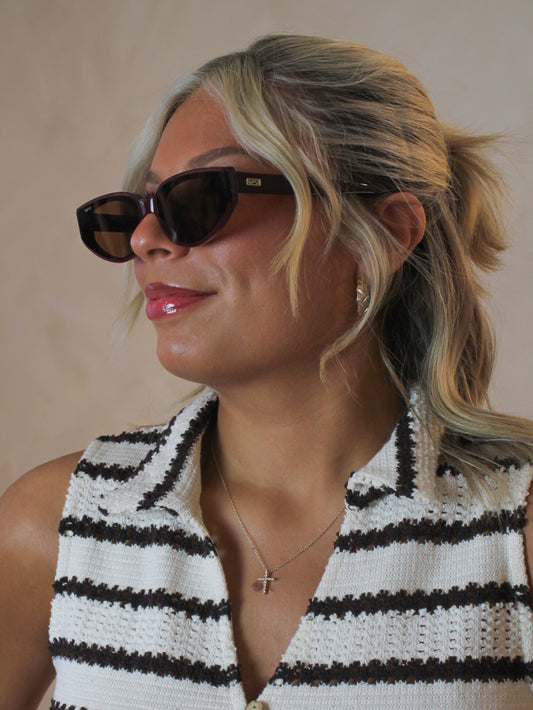 Woman wearing narrow cat eye sunglasses in a dark cherry red color with gold accessories and a striped knit top outfit