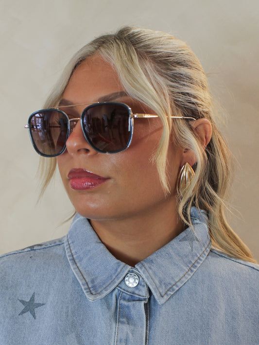 girl wearing blue, square aviator sunglasses with gold bars and hardware. Paired with a denim top for a cool girl outfit