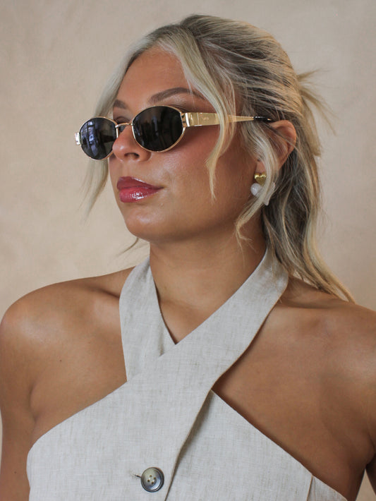 Woman wearing oval sunglasses with a gold frame and arms, black lenses and styled with a linen halter top 