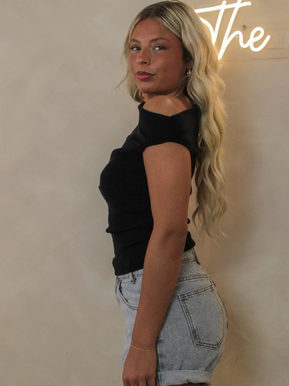 A woman shown from the side, wearing a black off-the-shoulder short-sleeve top and light-wash denim shorts.
