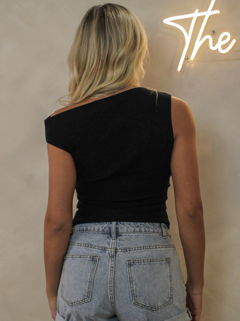 A woman viewed from behind wearing a black off-the-shoulder top and light-wash denim shorts.