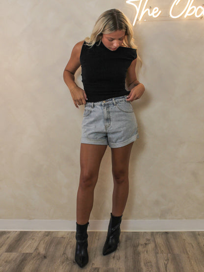 A woman modeling a black ribbed  off the shoulder top styled with light-wash denim shorts and black ankle boots